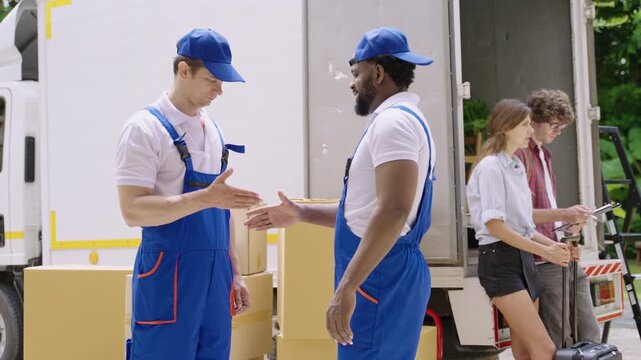 Happy professional movers in uniform standing talking and shaking hands after finishing unloading from a truck while a young couple of homeowners are checking and signing for delivery at a new home. 