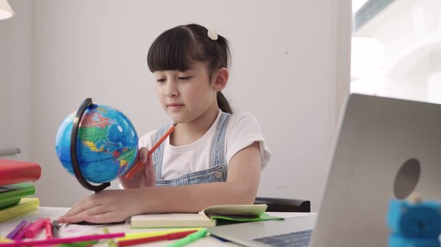 Cute Asian schoolgirl sitting at a desk using laptop computer for studying online learning via video calling and a remote teacher while having a geography lesson with a globe model in homeschool room.