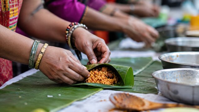 Hands of women preparing traditional food wrapped in banana leaves, with bowls and utensils visible on a table during a cultural cooking event