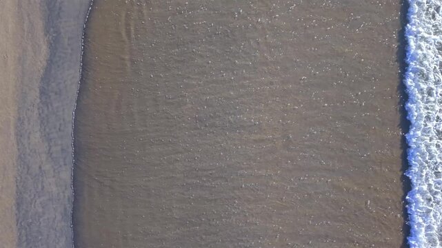 Minimalist top-down drone footage of gentle ocean waves washing onto a sandy beach, revealing natural textures, soft patterns and a peaceful coastal atmosphere.