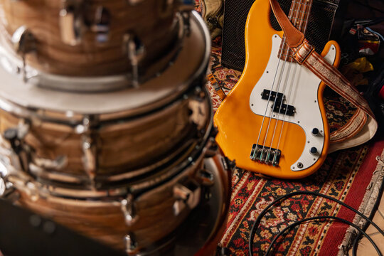 An orange four-string electric bass lies beside wood-grain drums and coiled cables on a patterned rug, rendered in warm vintage tones.