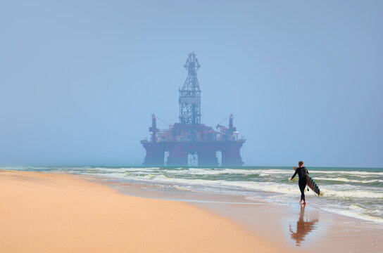 A male surfer walks on the beach with a surfboard in hand - Panoramic view of offshore jack up rig and offshore platform for oil and gas exploration and production - Namibia