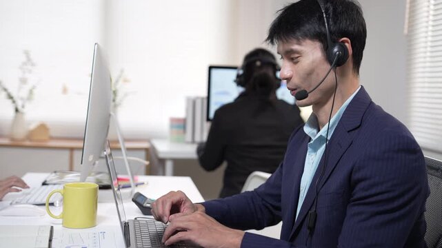 Asian man working in an office with a headset while looking at a computer