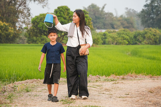 Asian mother holding small blue bucket over son head while walking together in green rice field, playful parent and child outdoors, happy rural family moment, traditional farm lifestyle and bonding