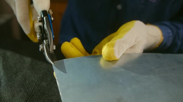 Skilled hands of a craftsman wearing protective gloves using heavy duty snips to accurately cut a piece of galvanized steel sheet in an industrial workshop, close up on the manufacturing process