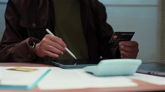 Person holding credit card, operating tablet, calculating personal finance budget expense payment online banking transaction securely indoor workspace managing stylus pen paperwork calculator desk.