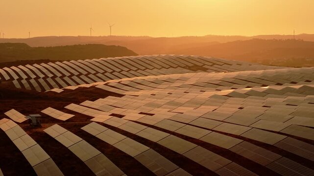  Aerial view of an extensive photovoltaic field glowing in warm sunset light, with wind turbines rising above distant hills