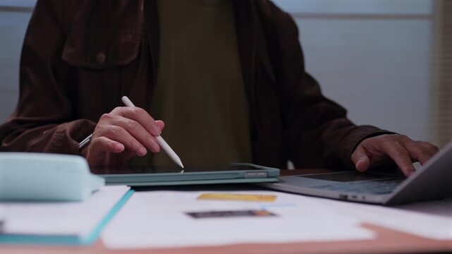 Person sitting desk holding stylus pen touching digital tablet screen typing laptop keyboard reviewing financial documents paperwork business project planning office environment indoor setting.
