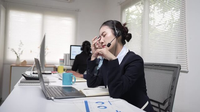 Exhausted Asian call center agent rubbing eyes from headache while working in office