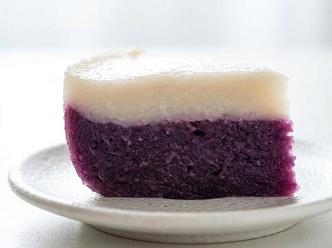 Slice of Indonesian traditional talam cake with two layers purple sweet potato bottom and white coconut milk top on white ceramic plate. Steamed glutinous rice flour dessert with vibrant natural color