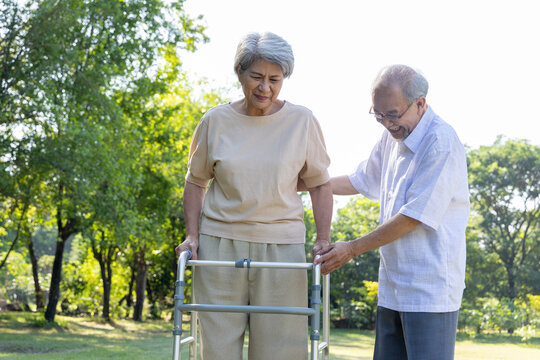 An Asian senior man lovingly helps his elderly wife walk using a mobility walker frame at an outdoor green park, providing physical support, encouragement, and compassionate caregiving