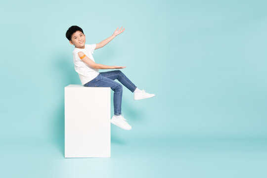 A happy Asian boy with a bandage on his arm sitting on a cube and presenting copy space, promoting child vaccination, healthcare campaign, and medical safety on light blue background