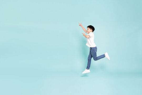 An energetic Asian boy jumping high and pointing up in studio, demonstrating joyful freedom, active movement, success concept, and child excitement on light blue background with copy space