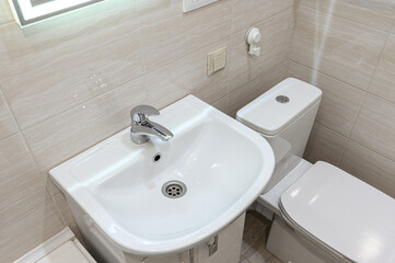 Fototapeta premium compact bathroom corner with a white ceramic sink and a chrome faucet. next to it stands white toilet, while beige textured tiles cover walls