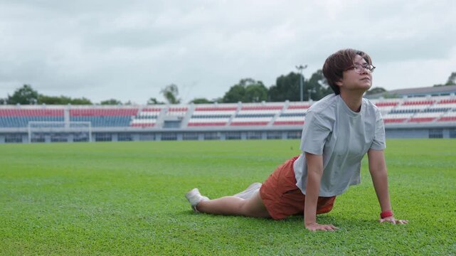 Authentic shot of an active Asian woman practicing a cobra stretch on a beautiful green soccer field. The empty stadium with colorful seats provides excellent copy space for sports backgrounds.