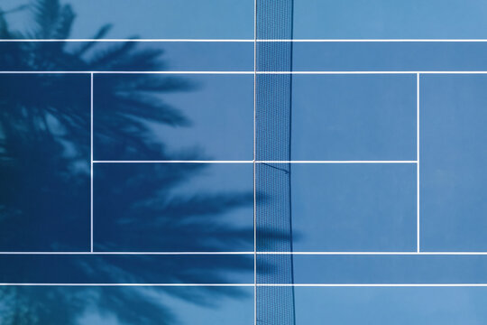 Aerial top down view of bright blue tennis court with white lines and net casting shadows, surrounded by palm tree shadows creating tropical atmosphere
