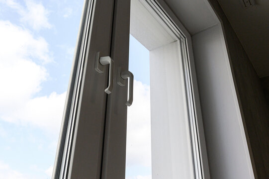 partially open PVC window sash with a sleek silver handle; the frame features clean white lines, and a bright sky with soft clouds is visible outside.