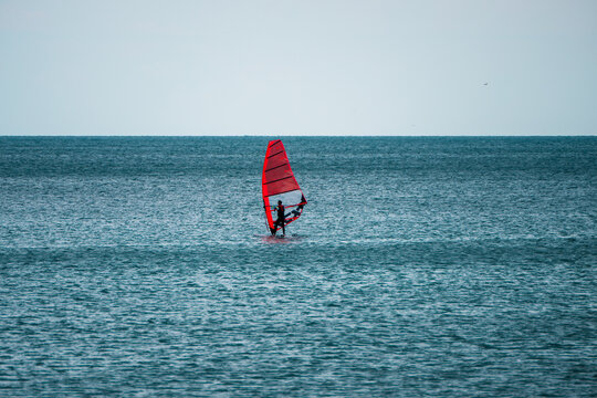 A windsurfer glides across the calm sea with an orange sail under an overcast sky.
