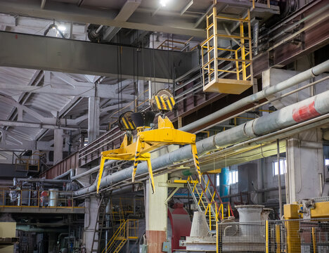 A large yellow crane grab tong mechanism hangs suspended from cables inside a heavy manufacturing factory workshop with pipes and steel support structures.