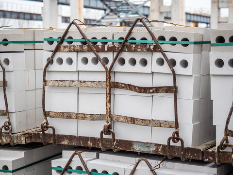 Stacks of white perforated concrete bricks are tightly bound with green plastic straps and secured inside rusty iron transport cages at an outdoor industrial yard.