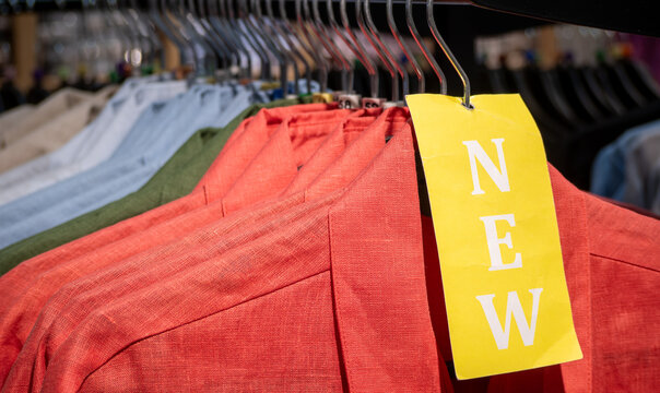 A yellow tag with the word "NEW" hanging on a metal hanger with coral-colored linen jackets among other colorful shirts on a store rack.