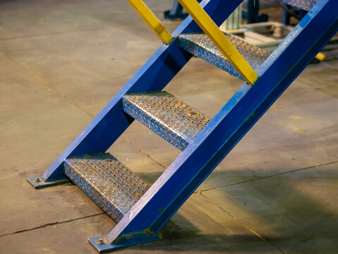 Heavy duty blue industrial metal staircase with diamond plate anti-slip steps and bright yellow handrails mounted on a concrete factory floor.