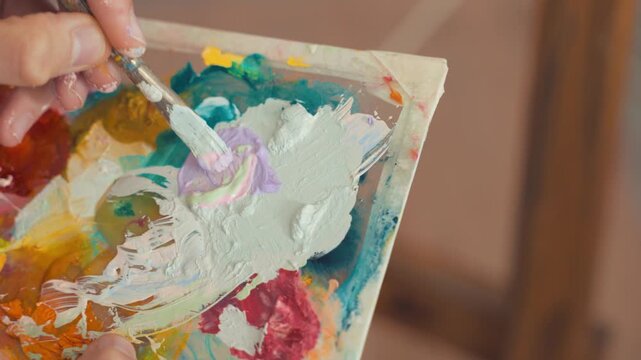 Artist carefully mixes light pastel oil paints on a wooden palette using a palette knife while focusing on creating a subtle color transition for the ongoing canvas painting inside the studio space.