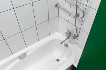 Fototapeta premium white acrylic bathtub with a chrome faucet, grab bars, and a shower hose. walls are tiled in white with dark grout, green partition is visible