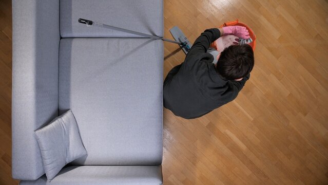 Person wearing pink rubber gloves dipping a mop head into an orange bucket of soapy water, preparing to clean the wooden parquet floor in a home setting