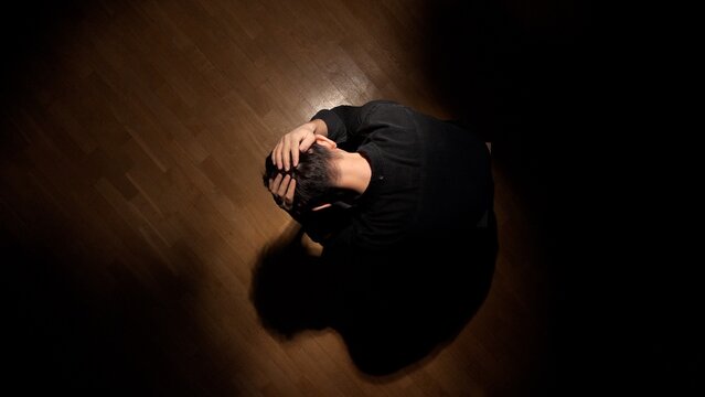 Depressed man with head in hands sitting alone on a wooden floor in a dark room, struggling with loneliness, anxiety, mental health issues, and stress