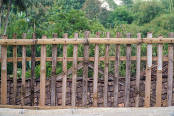 Fototapeta premium Rustic bamboo fence with weathered texture set against lush green forest backdrop and tiled roof. Natural, handmade construction ideal for rural or eco-friendly design projects.
