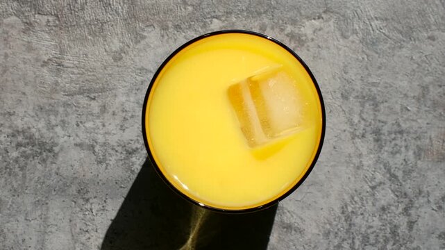 Top view hand placing large ice cube into glass of fresh orange juice on concrete background, perfect refreshing drink for sunny day, natural light strong shadows. Hand adding ice cube to orange juice