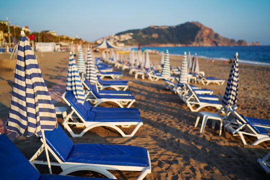 Rows of blue sunloungers under striped umbrellas line a sunny beach, creating a relaxing seaside resort vibe with calm water and distant rocky outcrop under clear skies.