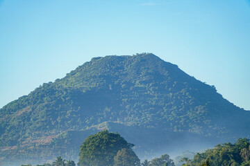 A majestic forested mountain peak under clear blue sky with misty foreground trees, showcasing natural beauty and serene landscape ideal for travel or nature themes, elegantly showcasing green. © Pernandi Imanuddin