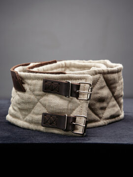 Quilted beige linen armor belt with double leather buckles displayed on a dark cloth surface.