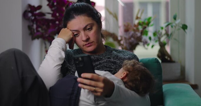 Mother scrolls smartphone while toddler reclines across her lap on living room sofa, balancing digital attention, childcare, multitasking and modern family life at home