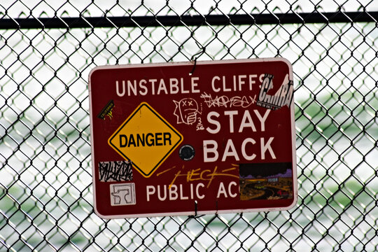 A graffiti-covered unstable cliffs warning sign hangs on a chain-link fence, advising visitors to stay back from the coastal bluff.