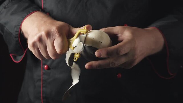A chef in a black coat with red accents continues to peel a black radish with a yellow peeler, exposing the inner flesh, while remaining focused on the task at hand in a well-lit kitchen environment