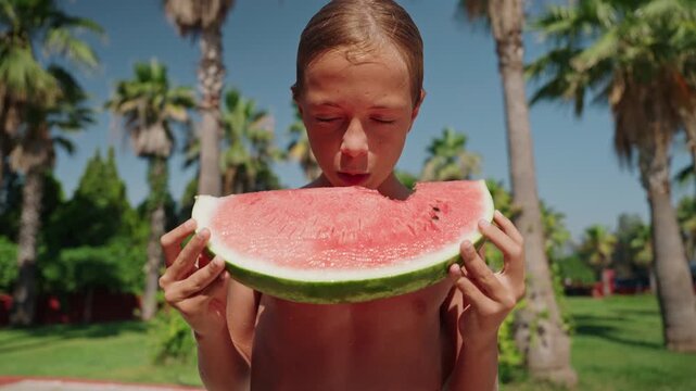 A happy young boy enjoys a large slice of fresh watermelon outdoors on a sunny summer day surrounded by palm trees and tropical greenery. Standing shirtless at a resort, he is about to take a bite of