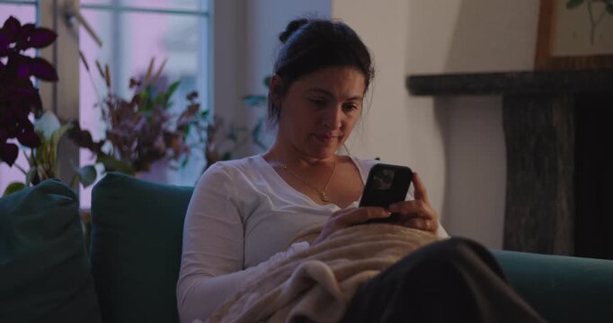 Woman scrolls smartphone while reclining on living room sofa under blanket at home, enjoying digital downtime, quiet relaxation, personal space and modern self-care