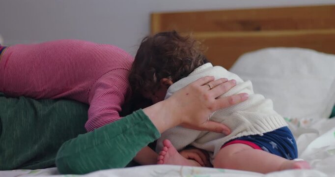 Toddler joins mother and older brother in group embrace on bedroom bed, expressing authentic maternal love, sibling affection, family unity and joyful togetherness