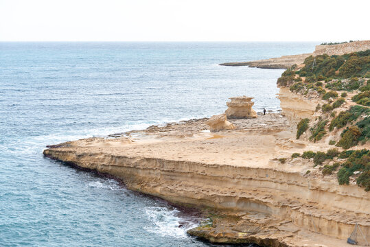 High angle view of a layered limestone cliff and rocky plateau by the Mediterranean sea featuring small rock stacks and a lone figure in the distance.