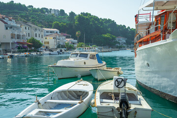 Small boats moored in a turquoise Mediterranean harbor with coastal houses and a green hill background. © JULIA