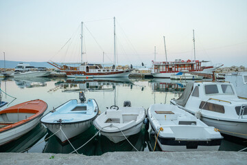 Fototapeta premium Various motorboats and traditional wooden yachts stay docked in a quiet marina harbor. The still water surface reflects the vessels under a soft twilight sky.