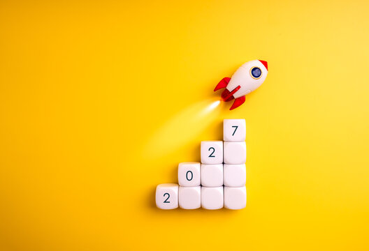 Rocket launches above white rising blocks marked 2027 on a vivid yellow background, symbolizing business growth, startup acceleration, strategic progress, ambitious goals and future success concept.