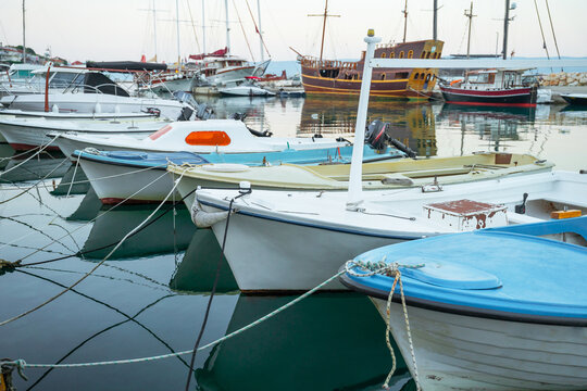 A row of small motorboats and skiffs docked in a peaceful marina with larger sailing vessels and coastal buildings visible in the background under soft light.