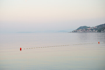 Calm sea surface with orange buoys and coastal town at dusk. Peaceful view of safety line leading to distant hills under a pastel sky. © JULIA