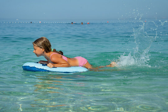 Young girl in a pink bikini lies on an inflatable board in clear turquoise ocean water, splashing and playing on a sunny summer day.