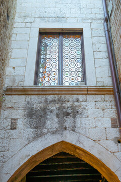 Vertical view of an ancient white stone wall featuring a pointed arch doorway and a window with circular glass panes and a brown metal drainpipe on the side.