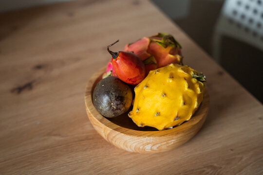 Exotic tropical fruits including yellow dragon fruit, pink pitaya, passion fruit, and tamarillo sit in a wooden bowl on a light-colored wooden table surface indoors.